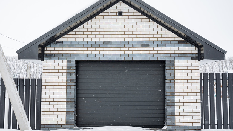 A grey garage door surrounded by a fence amid falling snow on a winter day