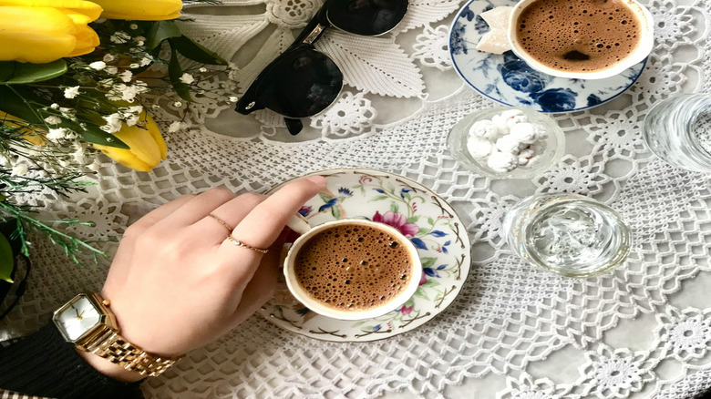 white crochet tablecloth with yellow tulips, sunglasses, coffee in a blue and white saucer and a hand