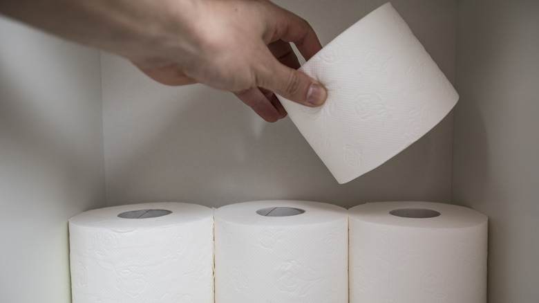 A person places a roll of toilet paper on top of three other rolls in a small white cabinet