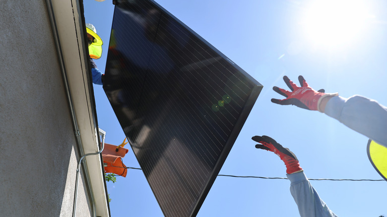 A large solar panel is lowered from the roof of a house to the outstretched arms of a person on the ground