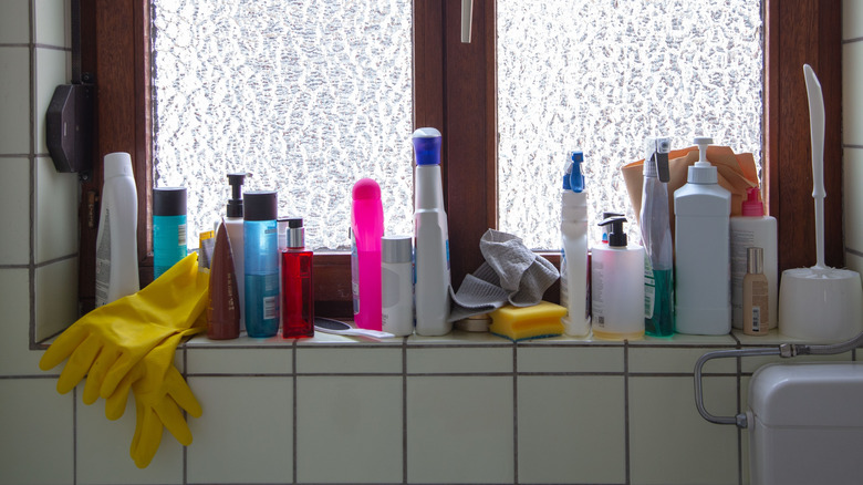A white tile bathroom has lots of products cluttering a windowsill