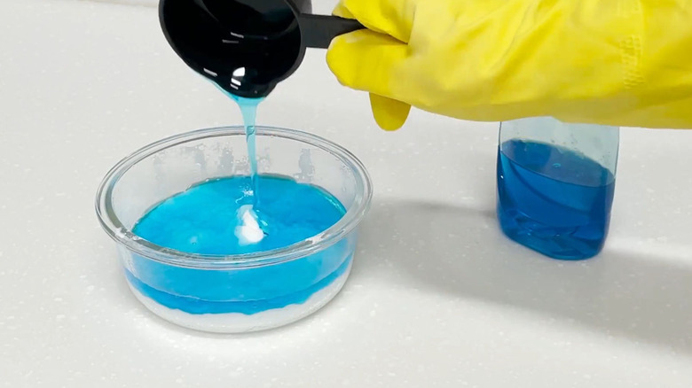 A gloved hand pours blue dish soap into a bowl of baking soda.