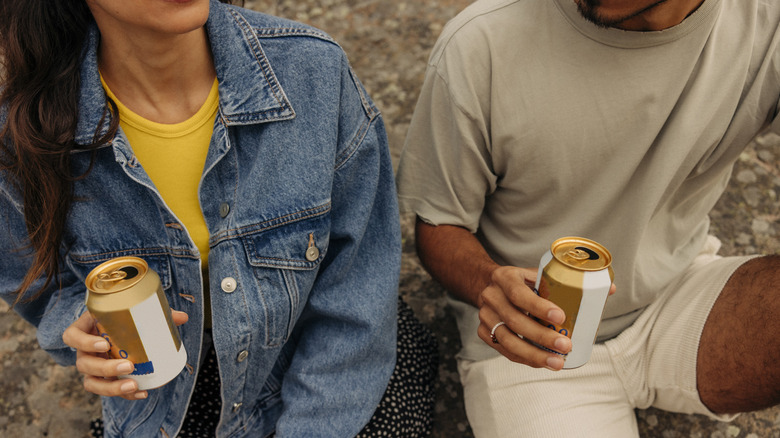 Two people hold soda cans as they drink.