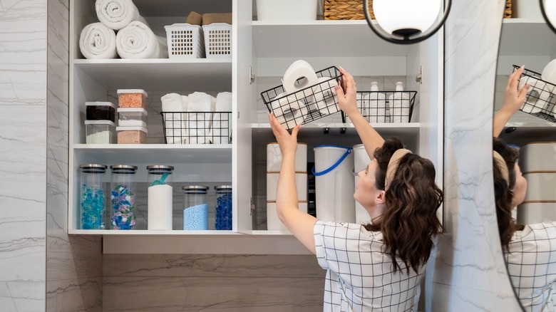 A woman places a basket of toilet paper on a deep upper shelf in an open cabinet