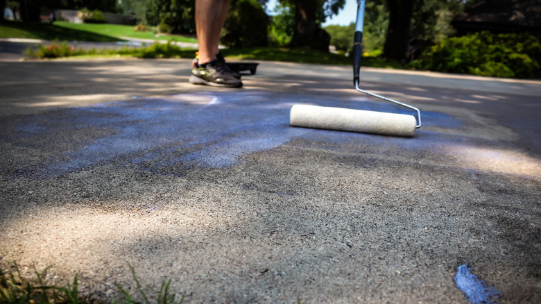 A person rolls sealant onto a concrete driveway using a paint roller.