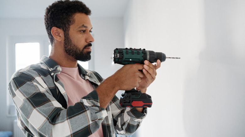 A man in flannel hold a drill pointed at a white wall
