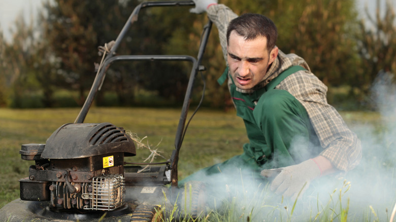 A man holds the handle of a lawnmower with smoke coming out of it.