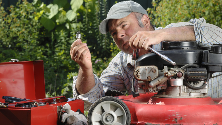 A man holds up a spark plug above a red gas lawn mower.