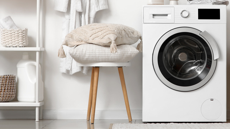 Modern washing machine with laundry basket and pillows on stool near white wall in bathroom