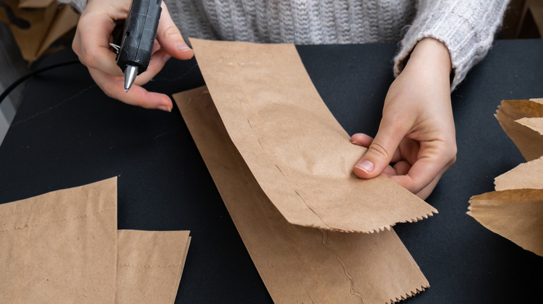 A woman's hands holding a hot glue gun and a brown paper bag on a table.