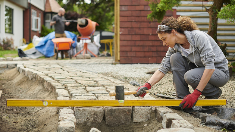 A person using a level while laying a brick path, while another person mixes concrete in the background