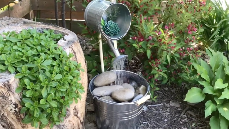 A homemade fountain using a watering can and a metal bucket, displayed in the corner of a garden.