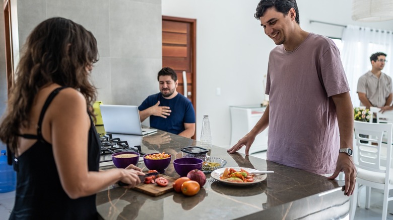 People standing, gathered around food on a stone countertop with a man seated by an open laptop.