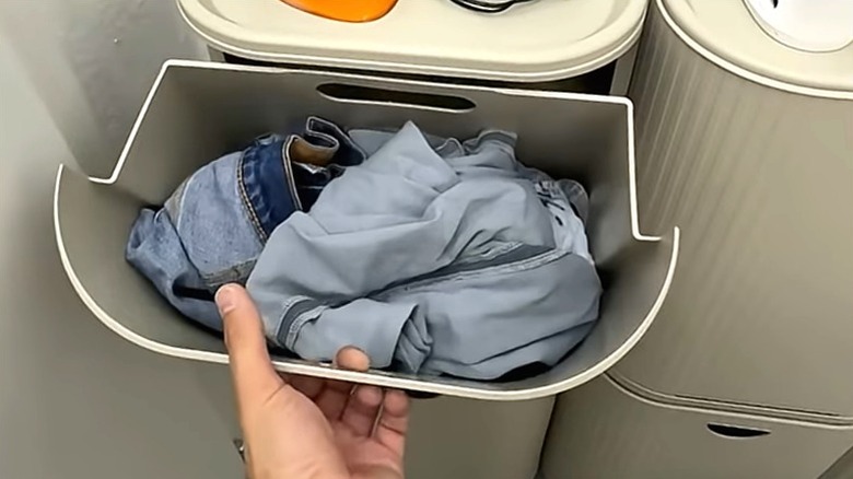 Person opening GÅRDSBUTIK bin on bathroom wall holding dirty laundry.