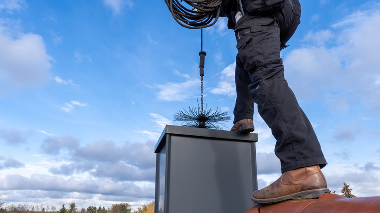 Chimney sweep stands on a roof, lowering their equipment down a chimney on a clear day