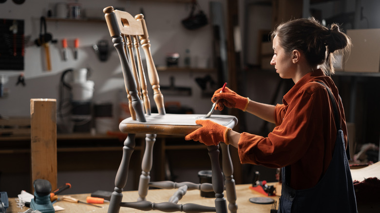 Woman painting a wooden chair grey wearing overalls and gloves