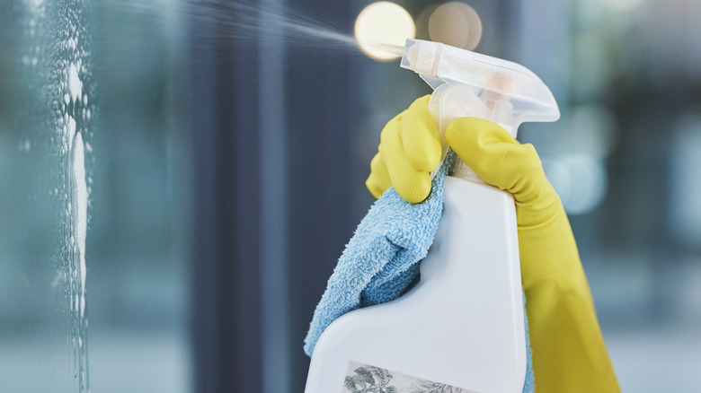 Gloved yellow hand spraying window cleaner on glass, rag in hand