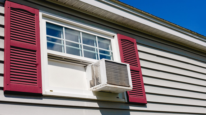 An exterior shot of a window air conditioner installed in a window in a house with light brown siding and red shutters