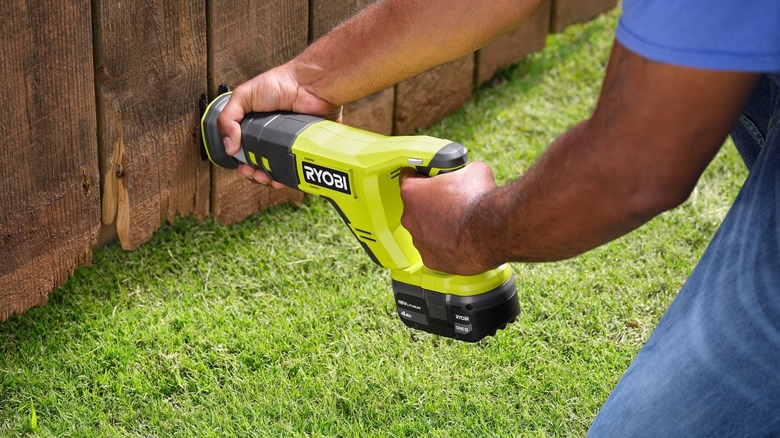 Person using a Ryobi reciprocating saw on a wooden fence with grass underneath