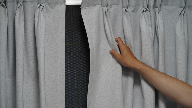 A single hand is opening the side of some pleated curtains over a dark window.