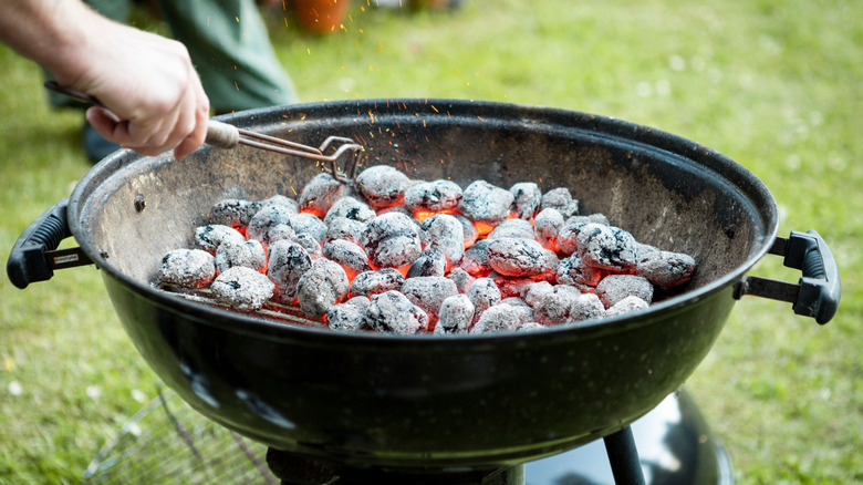 A closeup of person's hand with metal tool turning around burning charcoal in black barbecue in the yard