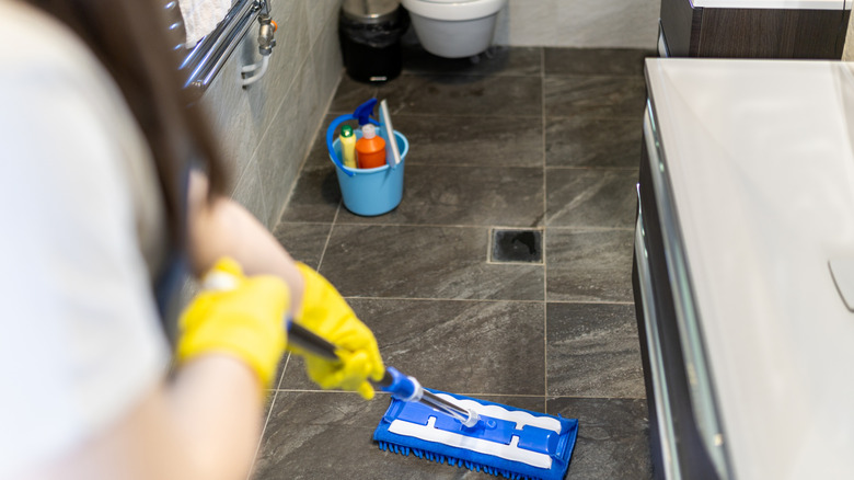 Person mopping in bathroom, their bucket of supplies nearby