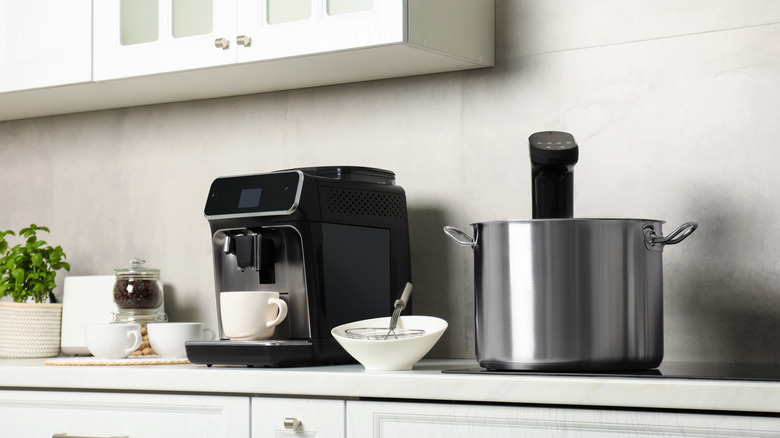 A kitchen with mugs, bowls, and appliances sitting on a white countertop, including a coffee machine and a pot with a sous-vide circulator