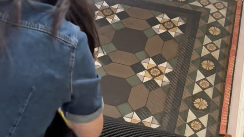 A woman pulling carpet in an entryway to reveal green and brown vintage tiles.
