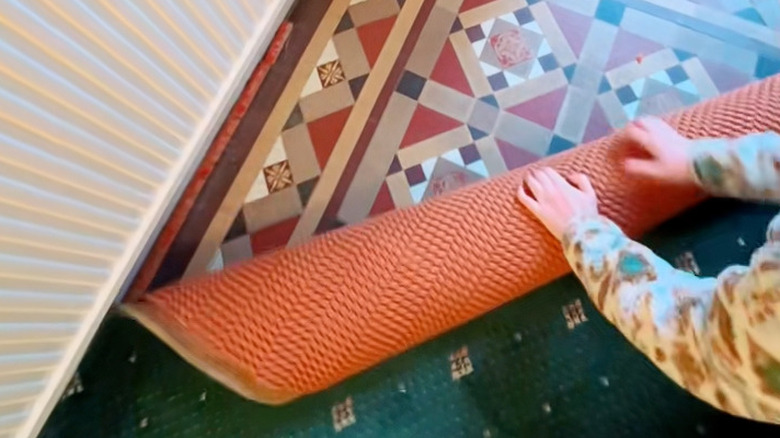 A woman peeling back a dark green carpet to reveal geometric tiling.