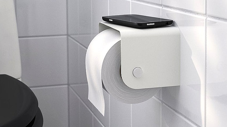 White metal toilet paper holder mounted on a white tile wall, with a black smartphone lying on the holder's shelf