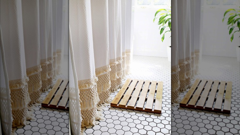 A long white shower curtain decorated with cream-colored fringe hangs in a white-tiled bathroom that also features a mat made of light wooden slats.