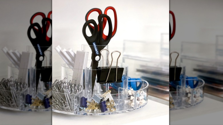 A clear plastic lazy susan with built-in compartments used to organize office supplies on a desk.