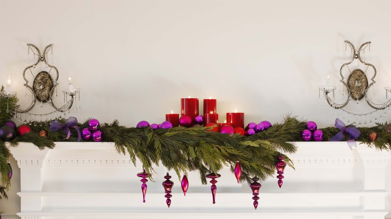 A green garland on the fireplace with red candles and red and purple ornaments with bows and a white background