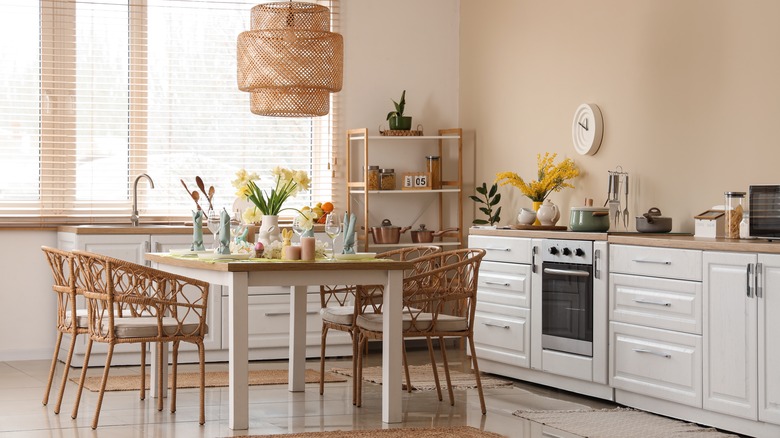 A kitchen with warm beige walls, white units, and a table and chairs with flowers and utensils on it.