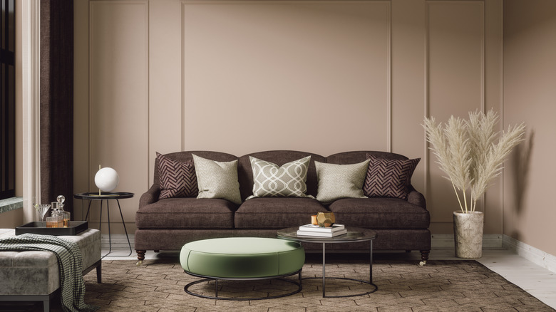 A brown sofa and green foot stool against beige walls with pampas grass in a vase in the corner.