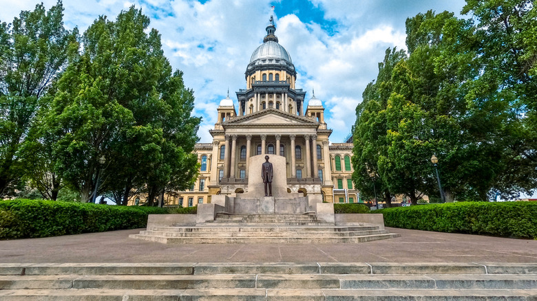 Front view of the Illinois State Capitol building and the bronze statue of Abraham Lincoln with cloudy blue skies overhead.
