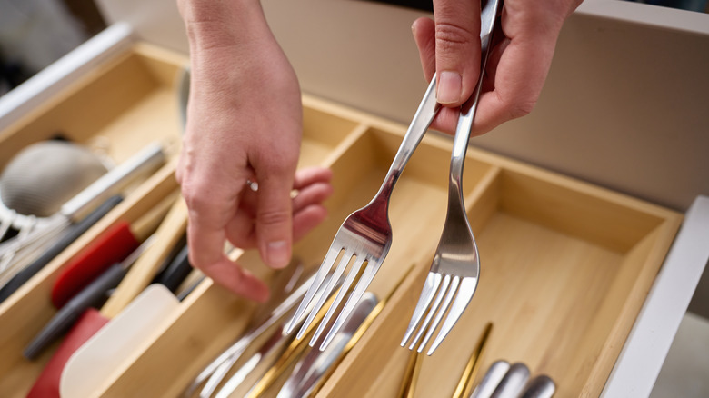 Two hands hold two stainless steel forks above a utensil drawer in the kitchen.