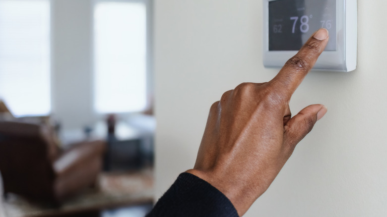 A man's hand pressing a touch screen thermostat with a living room in the background.