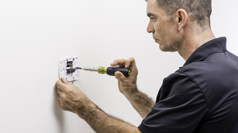A man in a dark blue work shirt using a screwdriver to remove a wall thermostat.