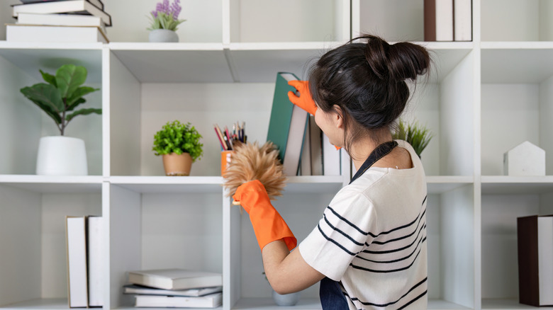 A person wearing orange rubber gloves using a duster on bookshelves.