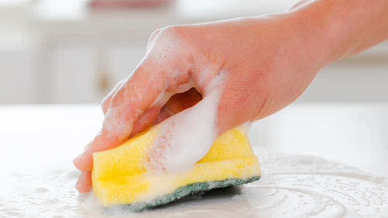 A close up of someone's hand cleaning a white counter with a yellow soapy sponge