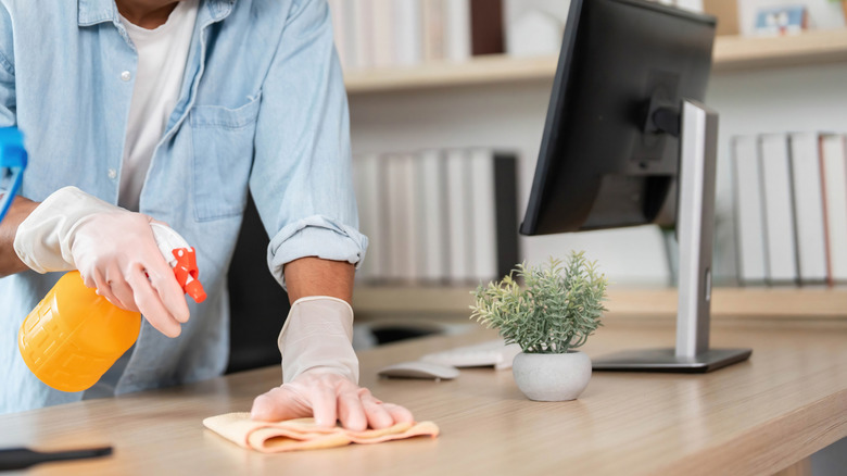 Someone wearing gloves and using a spray cleaner to wipe down a desk