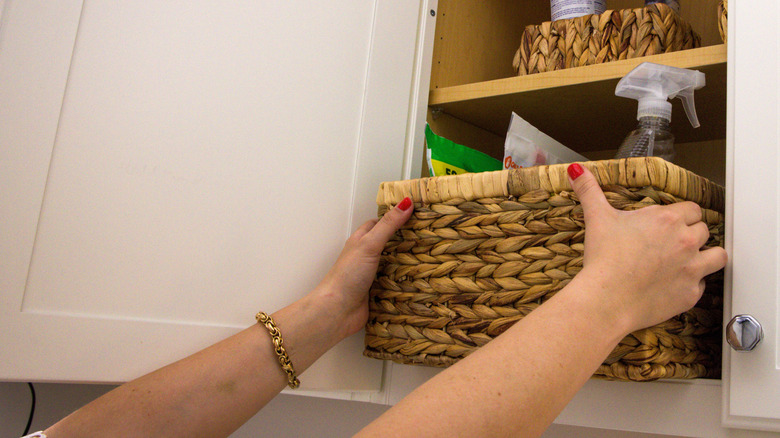 A person pulls a basket full of cleaning products out of the shelves in a laundry room storage cabinet.