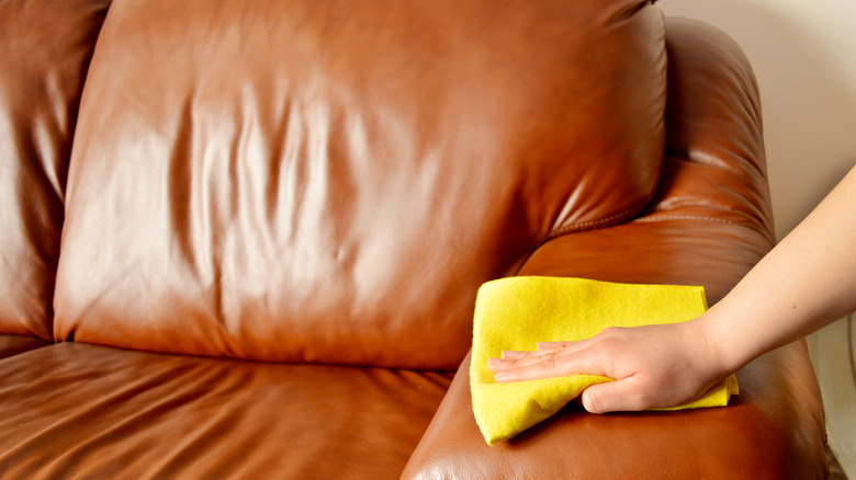 A person cleans a leather sofa using a yellow microfiber towel.