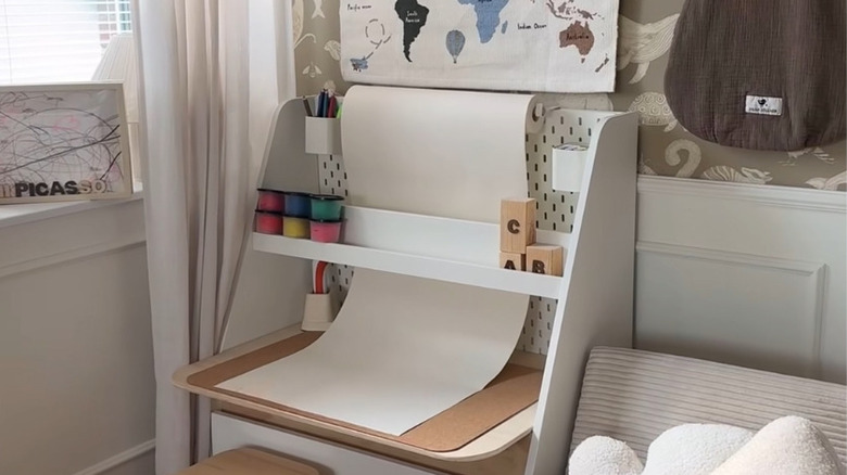 An IKEA pegboard is nestled into the back of a small bookcase, and an art paper roll rests on top, unfurled behind a shelf